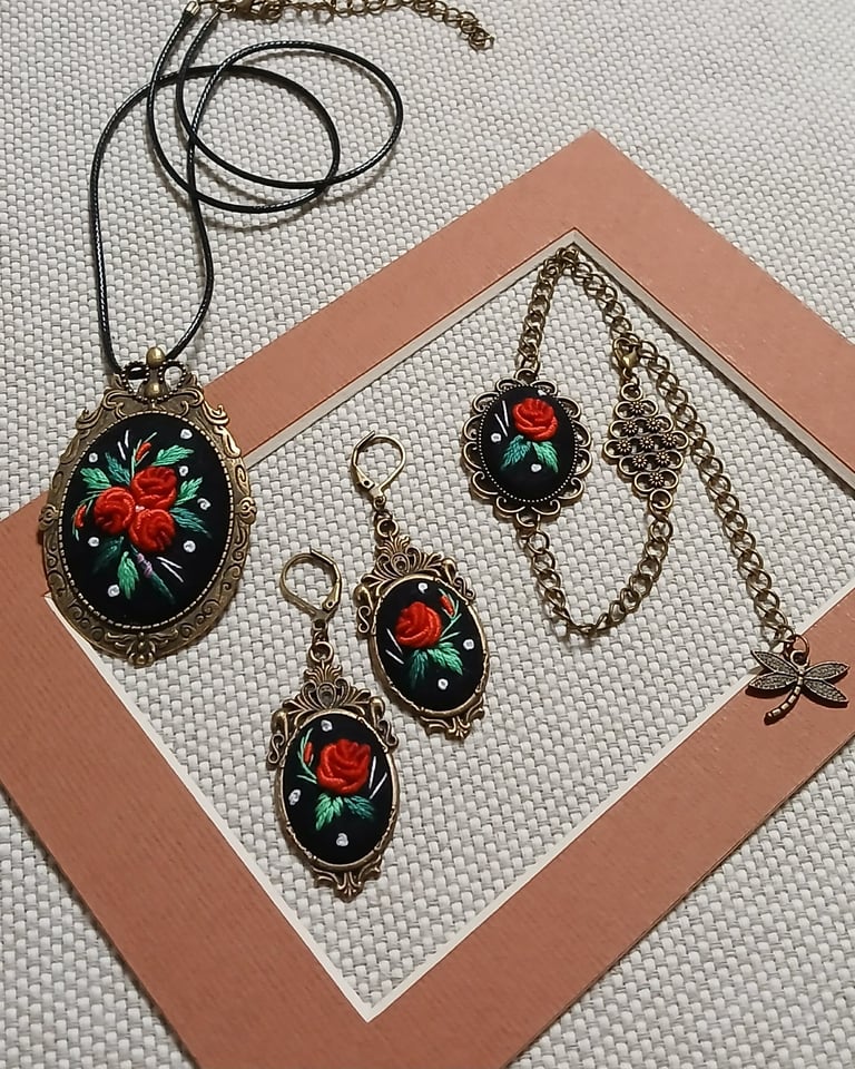 Collection of vintage-style jewelry featuring red rose designs on black enamel ovals, including a necklace, bracelet charm, and matching earrings with brass settings.