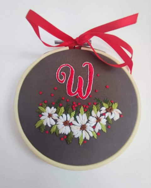 Embroidered hoop ornament with letter W, white daisies, and red ribbon bow on dark fabric