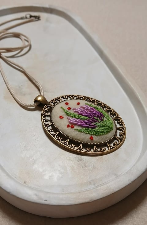 Ornate oval pendant with purple flower and red beads on silver chain, displayed on white ceramic dish