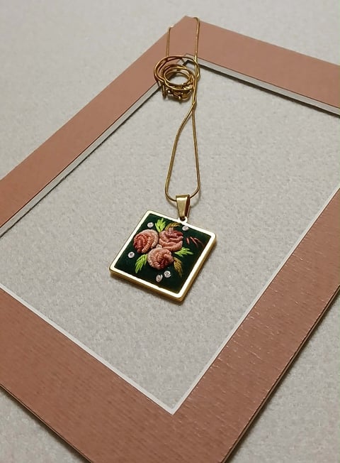 Gold chain necklace with square pendant featuring floral embroidery of pink roses on black background, displayed on white and rose-gold frame