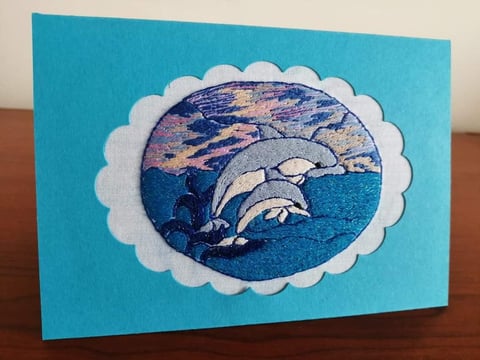 Handmade greeting card with blue background featuring a white scalloped frame around an embroidered dolphins jumping through ocean waves