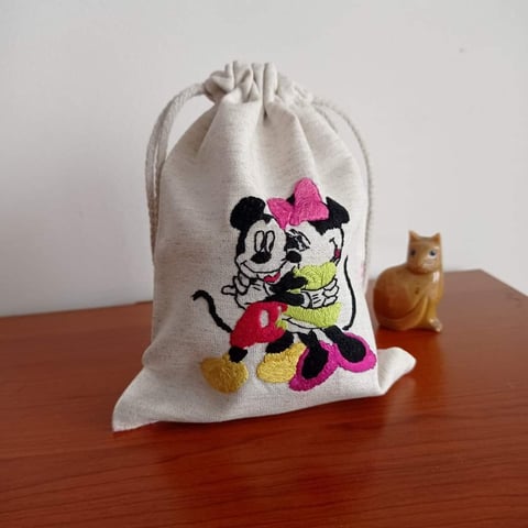 Beige cotton drawstring bag with embroidered Mickey and Minnie Mouse design on wooden surface next to small wooden cat figurine