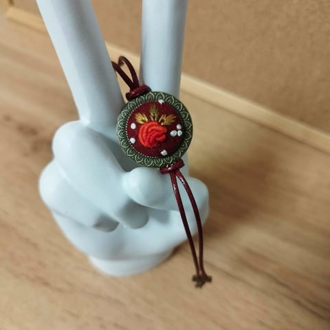 Decorative pendant on ring holder with red rose center, green beaded border, and burgundy strings
