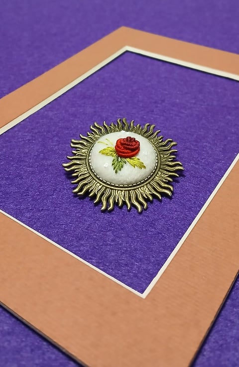 Ornate brass brooch with red rose on cream background mounted on purple mat with tan frame