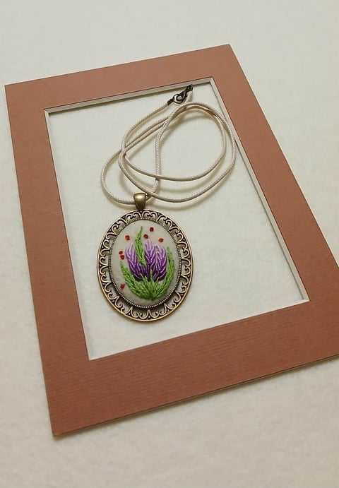 Ornate oval pendant necklace with embroidered purple flowers on beige cord displayed in brown wooden frame