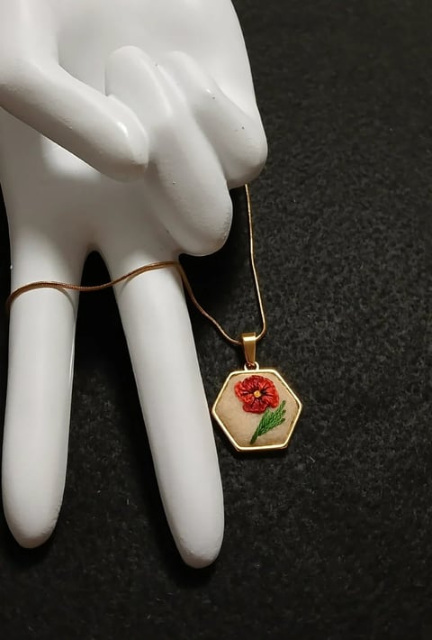 White mannequin hands displaying gold ring and hexagonal pendant necklace with red flower design on dark background