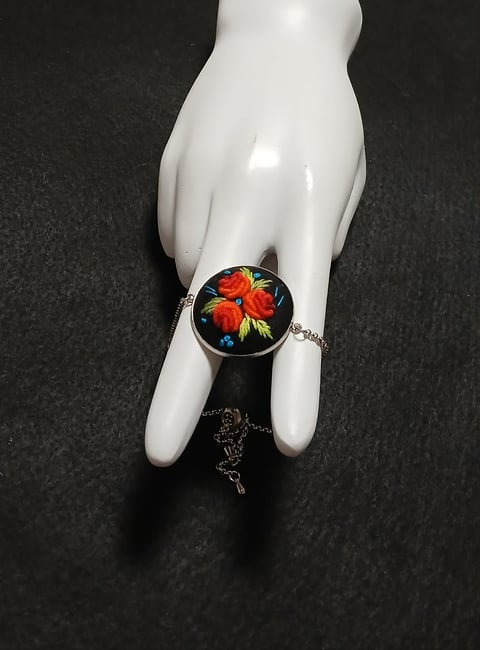 White mannequin hand wearing a silver bracelet with a round pendant featuring red flowers and blue accents on black background