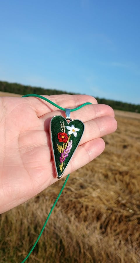 Hand holding a heart-shaped pendant with embroidered flowers on green ribbon in an open field
