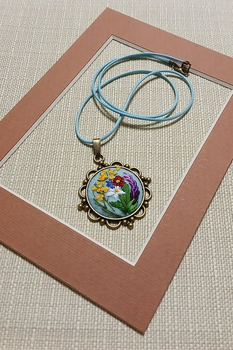 Ornate pendant necklace with colorful embroidered flowers on blue cord, displayed on a brown and cream matted background