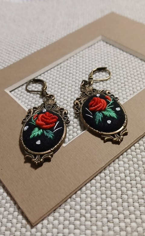 Pair of ornate brass earrings with embroidered red roses and green leaves on black fabric, displayed on beige card