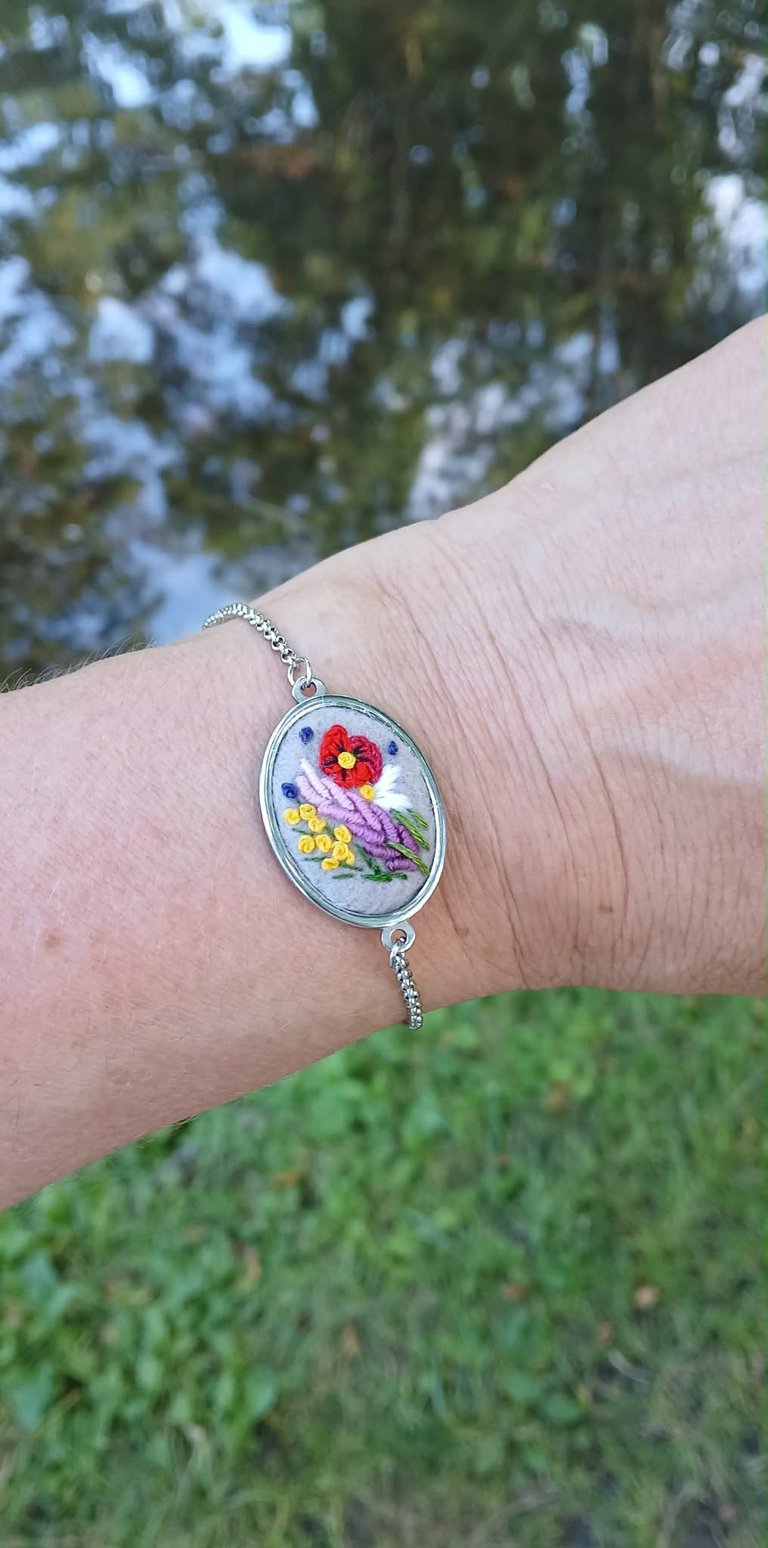 Hand wearing embroidered floral bracelet pendant with red, purple, and yellow flowers on wrist outdoors
