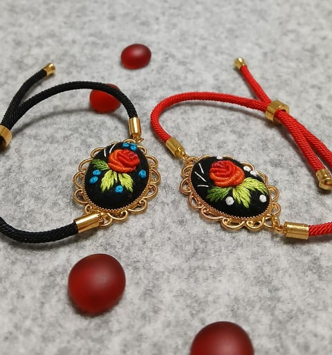 Two ornate bracelets with floral enamel pendants on black and red cords with brass accents, displayed with red beads