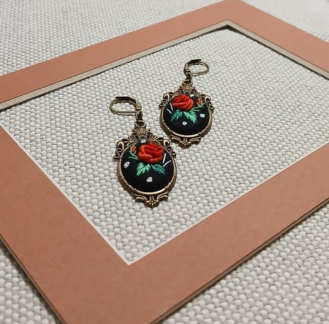 Pair of ornate vintage-style earrings with black enamel, red roses, and green gemstones displayed in a copper-rimmed box