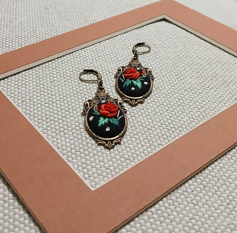 Pair of ornate vintage-style earrings with black enamel, red roses, and green gemstones displayed in a copper-rimmed box