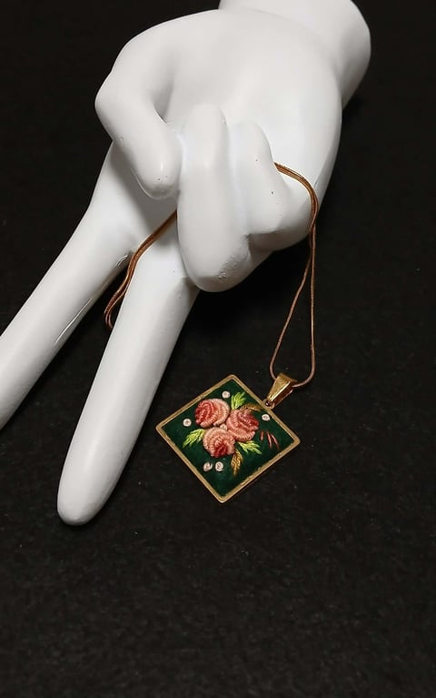White ceramic hand mannequin holding a square gold pendant with embroidered pink roses and green leaves on a copper chain