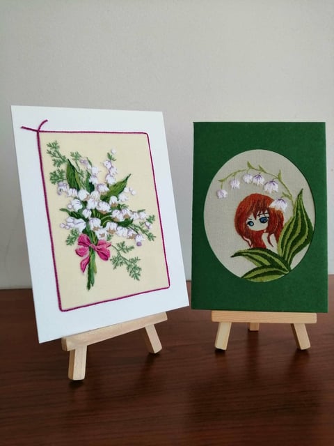 Two handmade greeting cards on wooden easels featuring embroidered floral designs with ribbons and flowers.