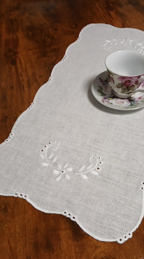 Delicate white linen tablecloth with eyelet embroidery on wooden surface, topped with floral teacup and saucer