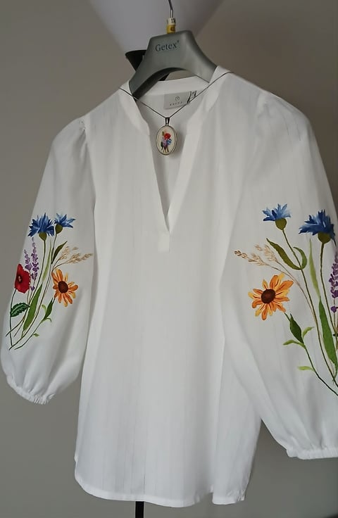 White embroidered blouse with colorful wildflower designs on puffy sleeves displayed on a dress form with pendant necklace