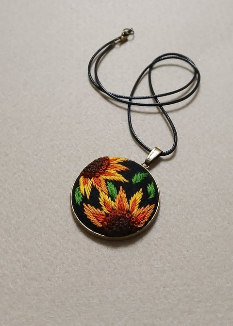 Pendant necklace with black cord featuring a circular metal-rimmed charm displaying embroidered sunflowers on black fabric