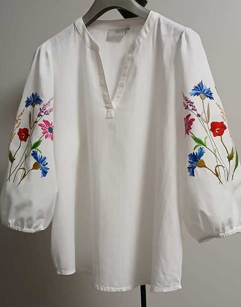 White linen blouse with colorful embroidered flowers on the sleeves, displayed on a hanger