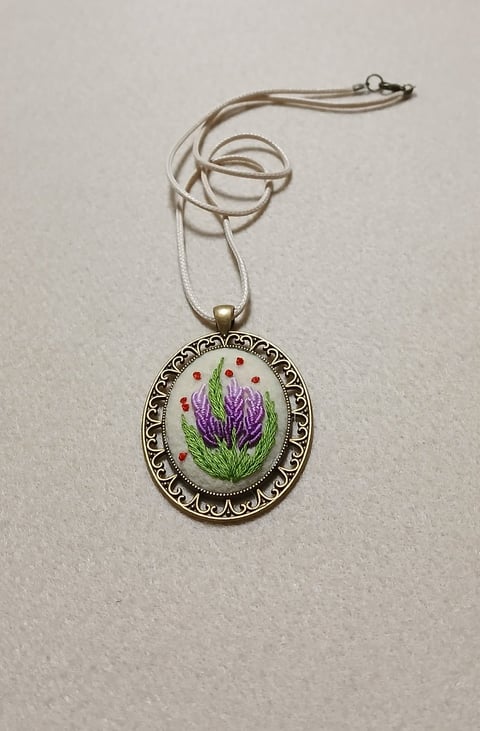 Silver chain necklace with ornate oval pendant featuring embroidered purple thistle flower with red beads on white background