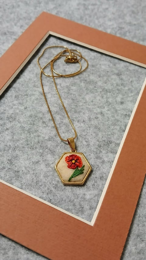 Gold hexagonal pendant necklace with embroidered red flower on cream background, displayed on coral and white mat
