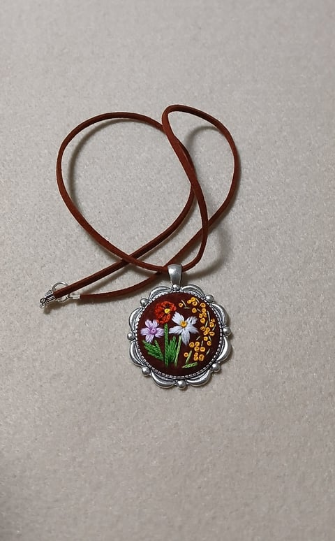 Brown leather cord necklace with ornate silver pendant featuring embroidered white and yellow flowers on dark background