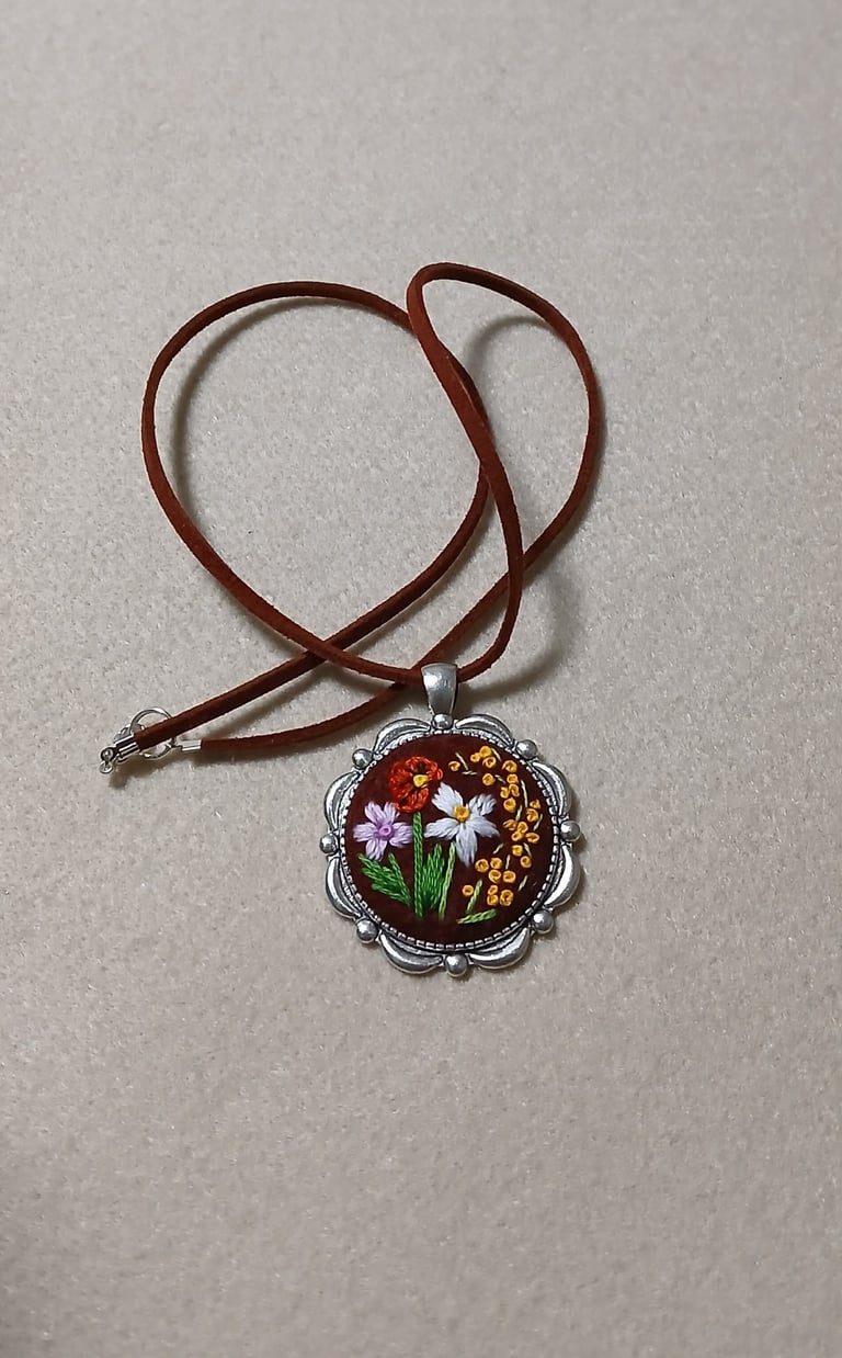 Brown leather cord necklace with ornate silver pendant featuring embroidered white and yellow flowers on dark background