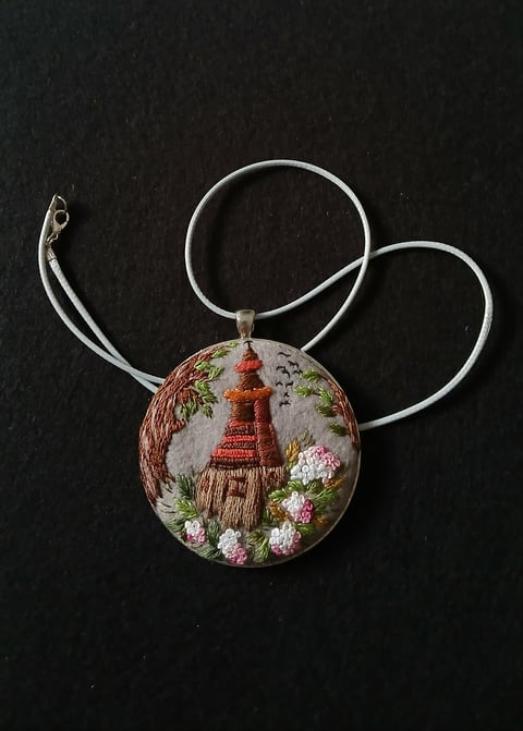 Embroidered pendant necklace with silver wire frame featuring a cottage and pink flowers on dark fabric background