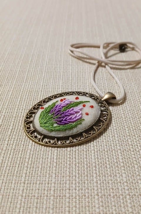 Hand-embroidered oval pendant with purple lavender flowers and green stems on white background, featuring ornate bronze frame and beige cord necklace