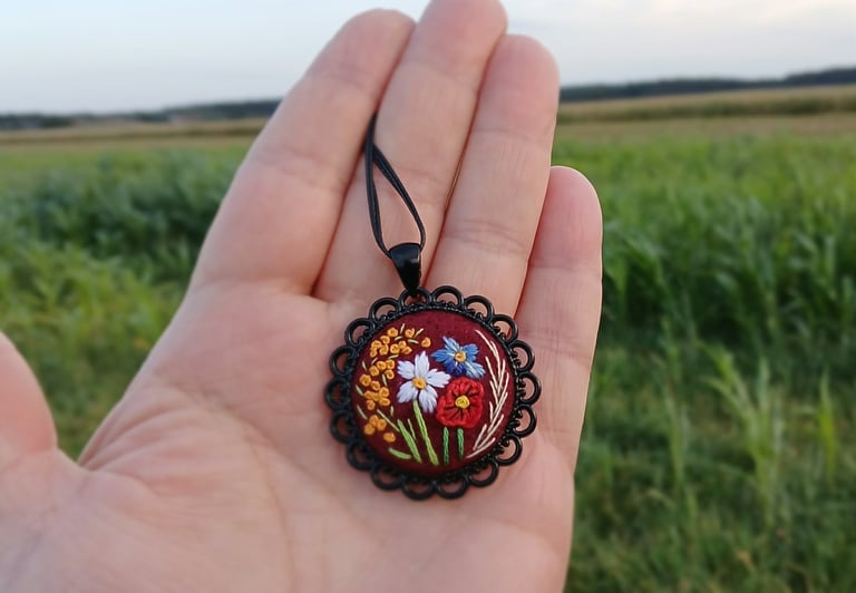 Hand holding a circular embroidered pendant with colorful flowers on dark fabric, displayed against a green field background