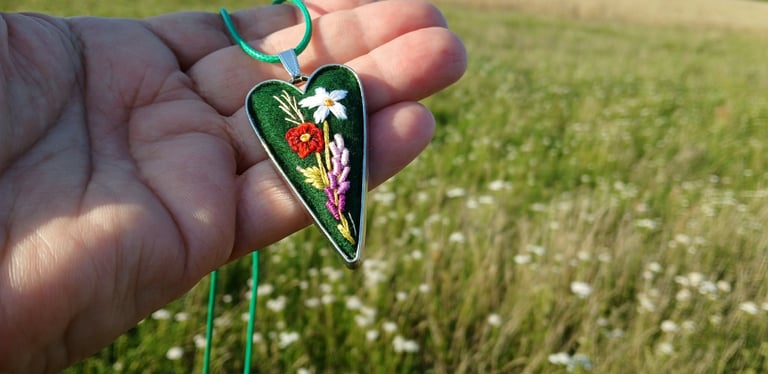Hand holding a green heart-shaped pendant with floral design on string, with wildflower field in background