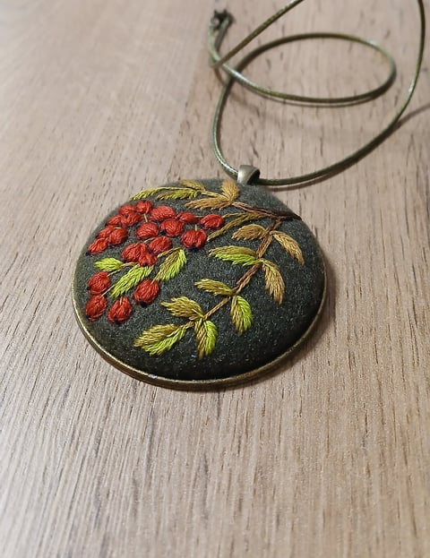 Embroidered pendant necklace with red berries and gold leaves on dark background, hanging from cord