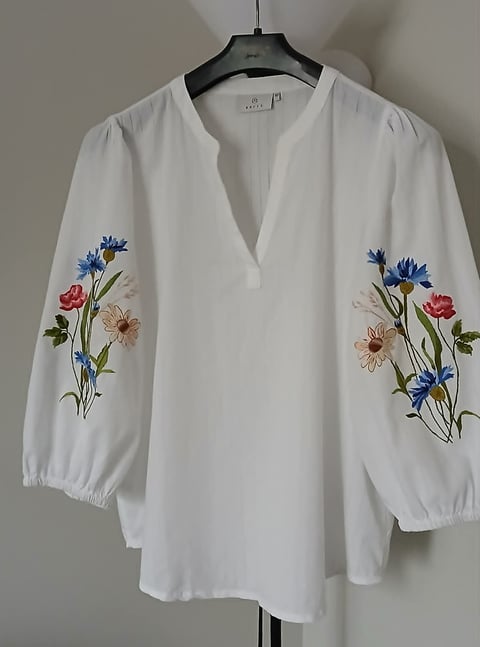 White blouse with embroidered flowers on sleeves, featuring blue cornflowers, red roses, and beige daisies, hanging on black hanger