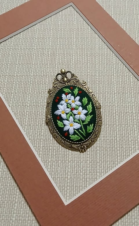 Vintage ornate oval brooch with white daisy flowers and green leaves on black enamel background, mounted in decorative brass frame