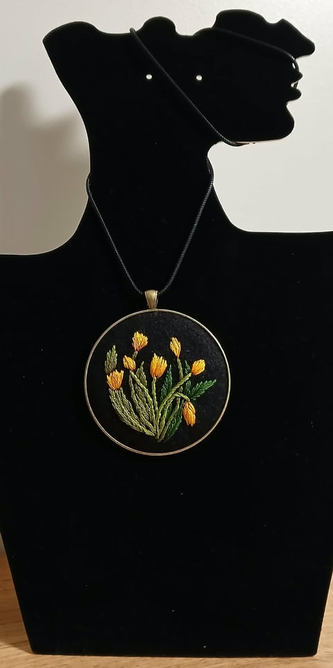 Black mannequin displaying a circular embroidered pendant necklace with yellow tulips on a dark background