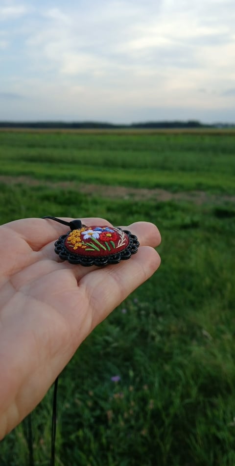 Hand holding a colorful embroidered ornament with floral design against an expansive green field and cloudy sky