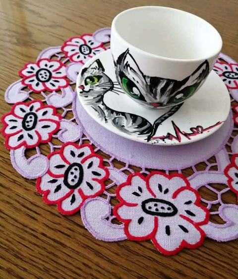 White teacup and saucer with black cat illustration on a pink and red floral doily on wooden table