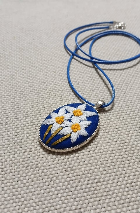 Blue oval pendant necklace with embroidered white and yellow daisies on blue cord, displayed on light fabric