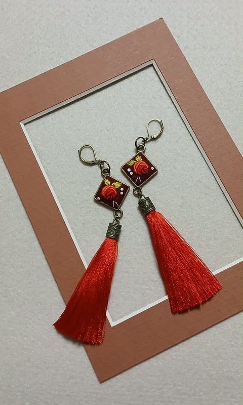 Pair of beaded earrings with red tassels and ornamental square pendants featuring floral designs, displayed on a tan framed white background