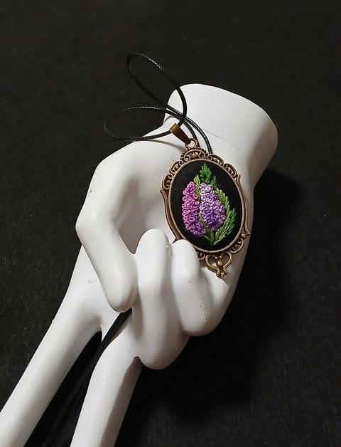 White skeletal hand holding ornate pendant necklace with purple lilac flower on black background
