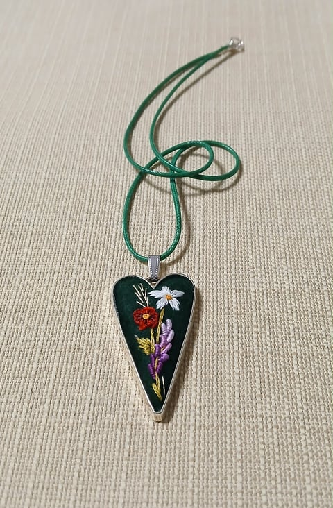 Heart-shaped black pendant with embroidered wildflowers hanging from a green twisted cord necklace
