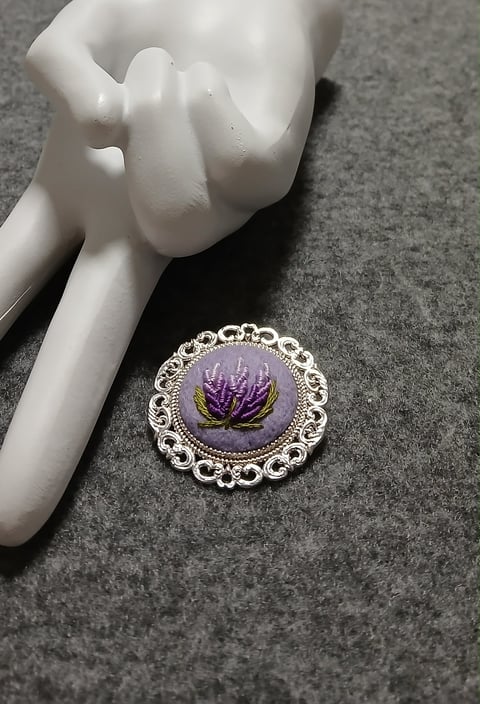 Ornate silver brooch with purple thistle embroidery on gray fabric next to white ceramic hand sculpture