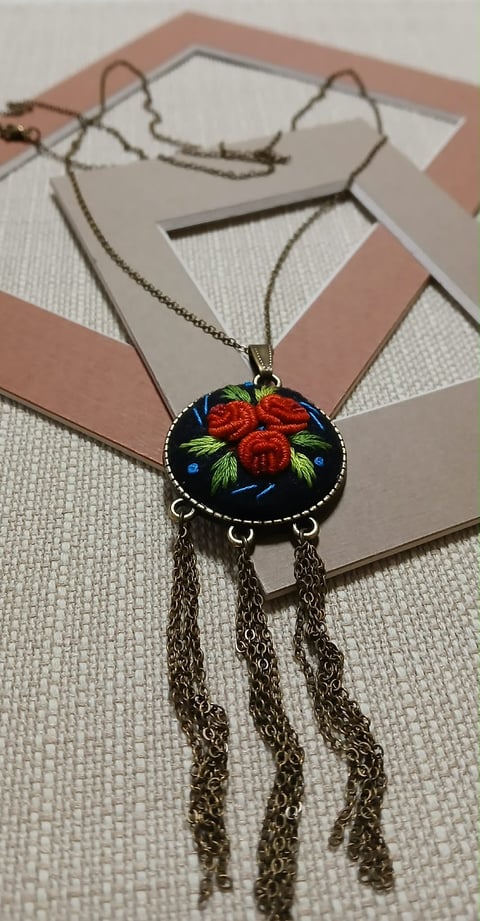 Embroidered pendant necklace with red roses on dark background, chain tassel details, displayed on kraft paper backdrop