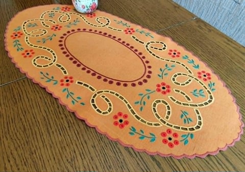 Decorative orange oval mat with hand-painted folk art design featuring floral patterns, colorful dots, and swirling gold trim on wooden floor