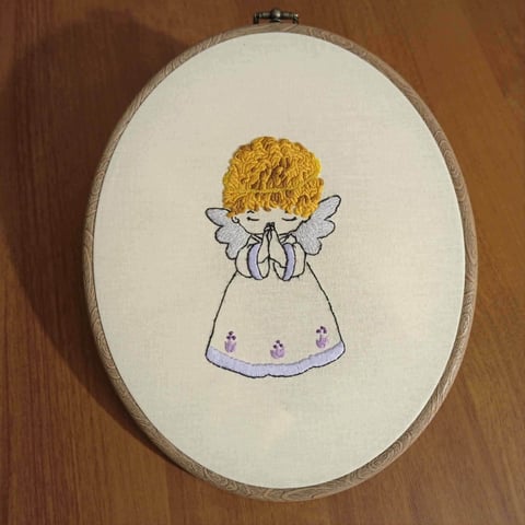 Embroidered artwork of a cute angel with yellow curly hair and purple dress with wings in an oval wooden hoop frame