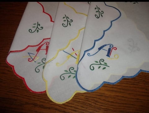 Three embroidered handkerchiefs with scalloped edges in red, yellow, and blue trim, featuring decorative stitched designs on white fabric