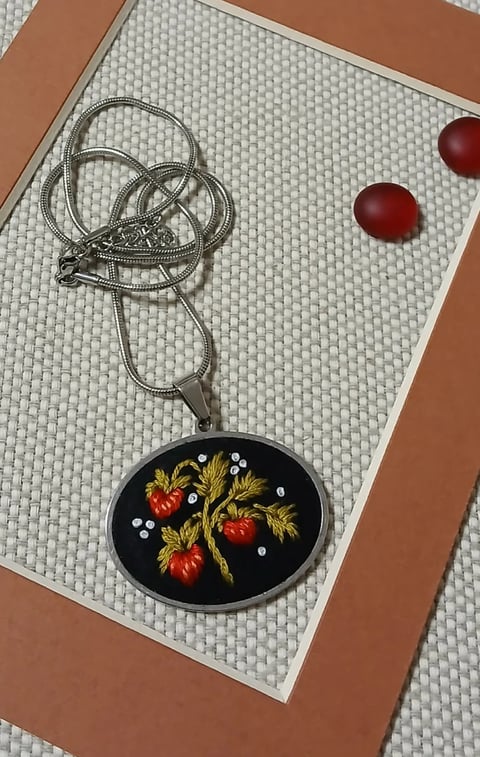 Black oval pendant necklace with embroidered red strawberries and gold leaves on white beaded background with brown frame